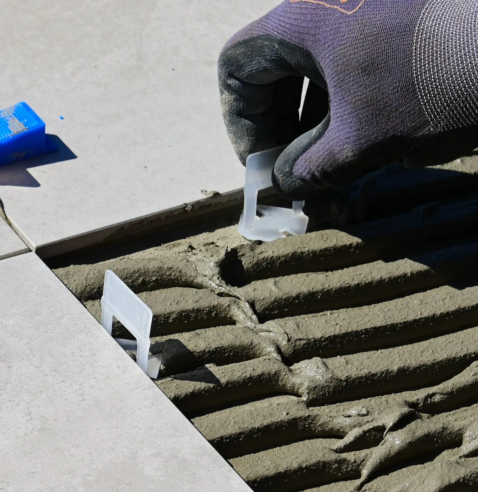 Hand im Arbeitshandschuh setzt Nivellierlaschen zwischen großformatige Platten, darunter gekämmter Fliesenkleber